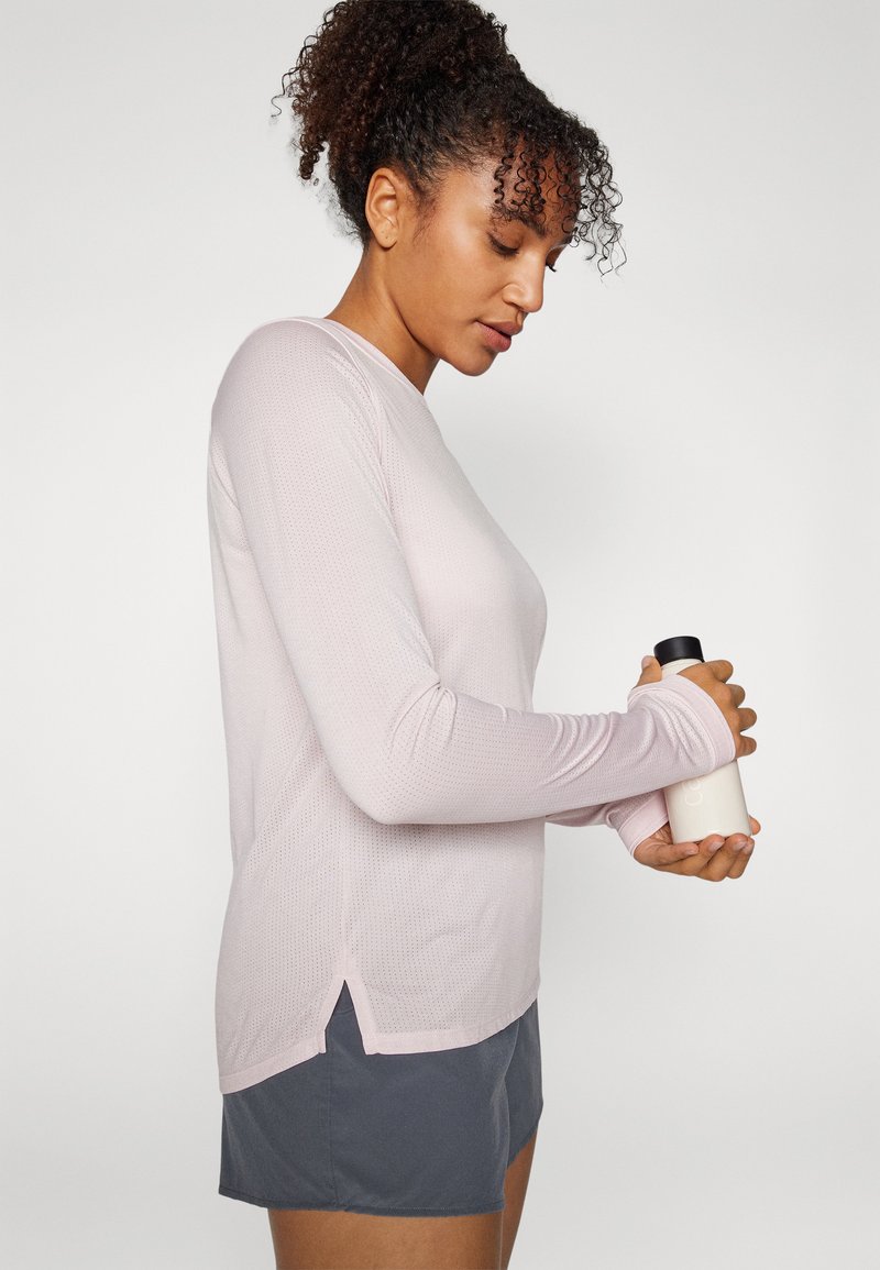 Helles rosa Langarmshirt mit strukturiertem Muster, Seitenschlitzen und Daumenlöchern, kombiniert mit dunklen Shorts; hält eine weiße Flasche mit schwarzem Deckel.
