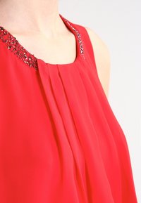 Top rosso senza maniche con pieghe e scollatura decorata con perline. Il tessuto ha una superficie liscia e il design è caratterizzato da una vestibilità ampia.