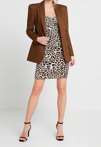 Ruskea räätälöity bleiseri leopardikuvioisen mekko-ihon päällä. Mekko on tiukka ja polven ylle ulottuva. Mustat remmikorot täydentävät asun.