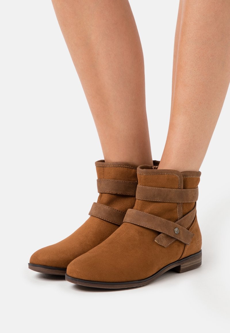 Bottes cheville en daim marron avec deux larges sangles et talons bas empilés portées sur des jambes nues sur fond blanc.