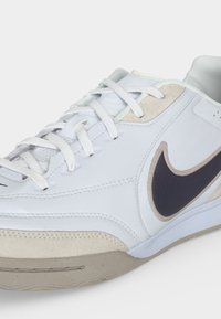 Λευκό και μπεζ αθλητικό παπούτσι Nike με μαύρο σήμα, λευκά κορδόνια και υφή στη σόλα, με γωνία που δείχνει τις λεπτομέρειες από το πλάι και από πάνω.