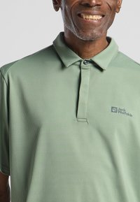Polo-shirt in een lichte groentint met een gestructureerde stof, een kraag, een knoopsluiting en een klein logo op de linkerkant van de borst.