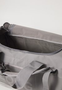 Saco de duffel de lona cinza com fecho na parte superior, alças grossas e detalhes em malha. O interior possui compartimentos para organização e uma impressão de logotipo discreta.