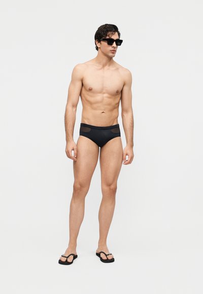 Άνδρας που φοράει μαύρο μαγιό τύπου βρακάκι, μαύρες σαγιονάρες και μαύρα γυαλιά ηλίου, στέκεται μπροστά σε απλό φωτεινό φόντο.