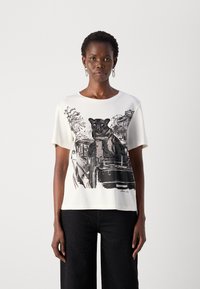 Vrouw in een wit T-shirt met een zwart-wit grafiek van een panter die op een stadsbus zit, gecombineerd met zwarte broek met kleine stippen.