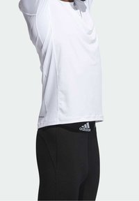 Vit sporttröja med meshdetaljer, korta ärmar och en avslappnad passform, i par med svarta leggings med en Adidas-logotyp på midjebandet.