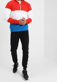 Uomo che indossa una felpa con cerniera rossa, bianca e blu, pantaloni jogger neri con strisce laterali bianche e sneaker nere, in piedi con le mani unite.