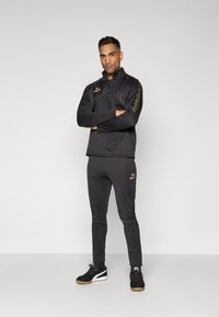 Melna sporta treniņtērps ar ceturtdaļas rāvējslēdzēju un slim-fit biksēm. Iekļautas zelta akcenti un zīmola emblēmas uz krūšu un sānu daļām. Melni sporta apavi.