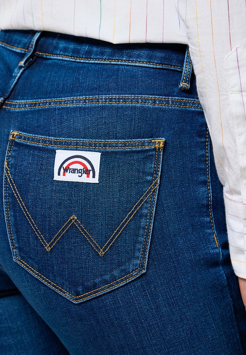 Niebieskie dżinsy charakteryzują się wyraźną tylną kieszenią z haftem w kształcie litery "W" oraz naszywką z logo Wrangler w czerwono-białym kolorze.