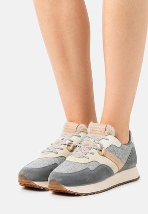 Piernas con zapatillas de deporte grises, beige y crema con tela texturizada, detalles de cuero, cordones estampados y suelas de goma sobre fondo blanco.