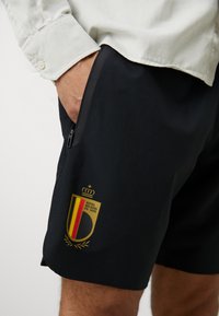 Melni sporta šorti ar rāvējslēdzēja kabatu un zelta, sarkanu un dzeltenu Beļģijas logo. Izgatavoti no viegla auduma, ar gludu tekstūru.