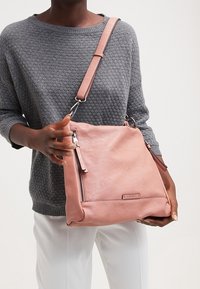 Sac à main en cuir rose avec une finition texturée, comportant une poche avec zip et une bandoulière amovible, porté sur un pull gris.