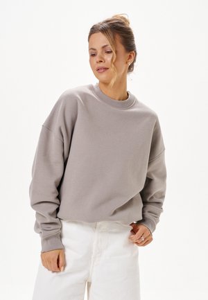 Vrouw met haar in een knot, gekleed in een losse grijze trui en witte broek, kijkt ontspannen naar beneden tegen een effen achtergrond.