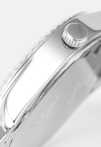 Silver wristwatch with a polished stainless steel case, featuring a textured crown and smooth bezel, emphasizing sleek, modern design.