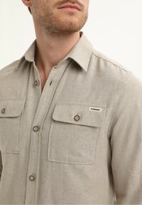 Camicia beige a maniche lunghe realizzata in tessuto testurizzato, con due tasche sul petto dotate di chiusura a bottone e un'etichetta sulla tasca destra.