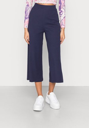 Vrouw die marineblauwe geribbelde wijde cropped broek draagt, witte platform sneakers en een kleurrijk langarmshirt met abstracte patronen.