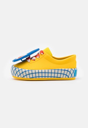Zapatilla deportiva amarillo brillante con cordones rojos y blancos, suela con patrón de cuadrícula azul y blanca, y un objeto circular azul unido en la parte superior.