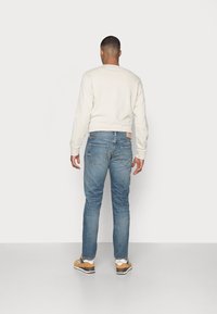 Sudadera gris clara con cuello redondo, combinada con jeans azules medios que presentan un aspecto desvanecido y bolsillos traseros. Zapatillas marrones con acentos blancos.
