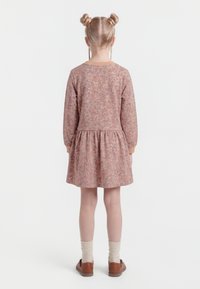 Bambina con capelli biondi raccolti in due chignon, indossa un vestito rosa a fiori a maniche lunghe, calzini crema e scarpe marroni, in piedi di spalle.