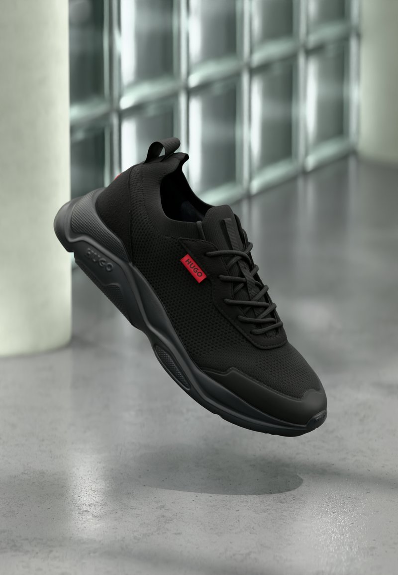 Baskets athlétiques noires en tissu mesh, semelle noire, laçage à l'avant, étiquette rouge "HUGO" sur le côté, présentées sur un fond intérieur moderne flou.