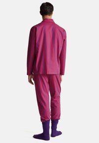 Muž stojící zády, oblečený v červeném a fialovém dlouhém tričku s vertikálními pruhy a kalhotách, s fialovými ponožkami na bílém pozadí.