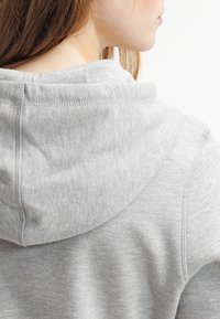 Sudadera con capucha de color gris claro, con un tejido suave y texturizado. Cuenta con una capucha con cordón y detalles de costuras visibles en los bordes.