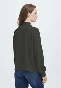 Femme aux longs cheveux bruns portant un blouse noire à manches longues avec un col et des poignets à volants, associée à un jean en denim bleu, se tenant de profil.