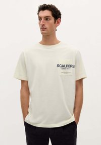 Tricou scurt cu mânecă din bumbac, de culoare crem, cu un buzunar pe pieptul stâng pe care este inscripționat textul "SCALPERS COMPANY" în albastru marin.