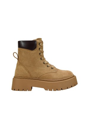 Botas de tobillo de ante marrón con suela gruesa y robusta, punta redonda y detalles en cuero marrón. Presentan ojales y una tira en la parte trasera.
