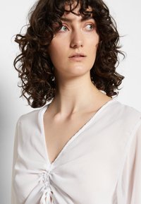 Blusa branca, texturada, com um decote em V profundo e detalhe franzido ao centro, apresentando mangas longas e um tecido leve e suave.