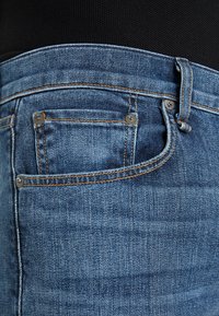 Los jeans de mezclilla azul presentan un diseño clásico de cinco bolsillos, costuras naranjas en contraste y un pequeño bolsillo adicional en la parte delantera.