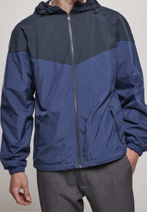 Mann trägt eine blau-schwarze Kapuzenjacke mit Reißverschluss, elastischen Bündchen und seitlichen Reißverschlusstaschen, steht mit einer Hand in der Tasche.