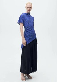 Top blu stripe con orlo asimmetrico, abbinato a una gonna plissettata nera. La texture del tessuto è liscia e sandali piatti completano il look.