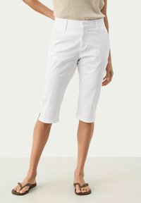 Shorts blancs arrivant aux genoux avec des fentes latérales, design ajusté et texture lisse. Assortis à des tongs marron et un haut en tricot beige.