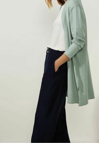 Ljusgrön lång cardigan med avspänd passform, bärs över en vit topp och mörk marinblå vida byxor. Har knappdetaljer vid midjan.