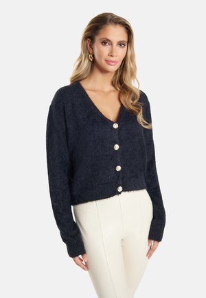 Svart fluffig cardigan med v-ringad hals, prydd med fem runda knappar, långa ärmar och en kort design. Bärs över krämfärgade byxor.