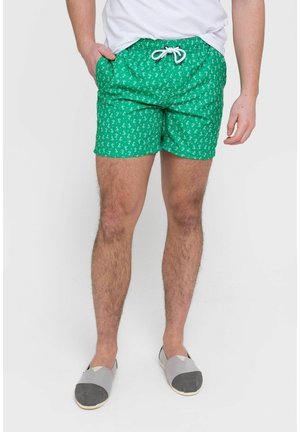 Hombre con pantalones de baño verdes con estampado y cordón blanco, camiseta blanca y zapatos sin cordones grises, de pie sobre un fondo liso.