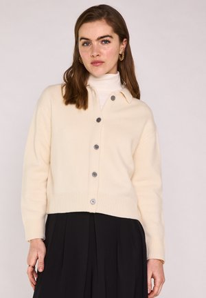 Jeune femme aux cheveux bruns mi-longs portant un cardigan crème boutonné sur un col roulé blanc et une jupe plissée noire sur fond neutre.