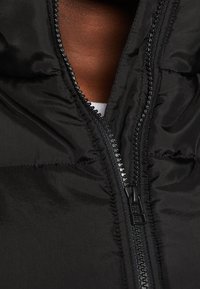 Musta vaihdettavalla takilla, jossa on sileä kankaan rakenne, vetoketjukiinnitys ja korkea kaulus. Vetoketjussa on mustat helmiäiskoristeet.