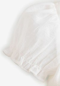 Tissu blanc et léger avec des bords de manches froncés doux et une texture subtile. Présente de délicats volants à l'ourlet des manches.