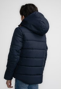 Marineblauwe pufferjack met een capuchon, voorzien van horizontaal doorgestikte secties. Het heeft een gladde textuur en een iets langere achterkant.