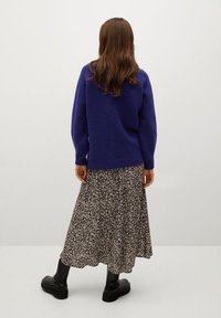 Donna con i lunghi capelli castani, che indossa un maglione viola, una gonna midi a fiori e stivaletti neri, in piedi contro un muro semplice, voltata di spalle.