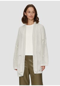Leichte, offen gestrickte weiße Strickjacke mit lockerer Passform, die mit komplizierten Mustern und überschnittenen Schultern gestaltet ist, über einem weißen Oberteil getragen.