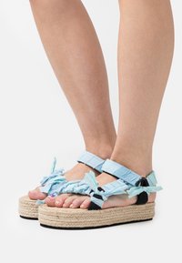 Ljusblå tygsandaler med volangdetaljer, platt vävd jutesula och blå- och svartrandiga remmar för ett säkert grepp.