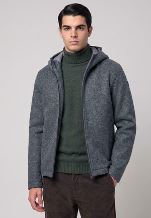 Giovane uomo che indossa una felpa grigia con zip, un maglione verde a collo alto e pantaloni marroni, in piedi davanti a uno sfondo chiaro e semplice.