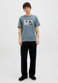 Grå bomulds t-shirt med grafisk tryk, der viser palmer, parret med sorte bukser og sorte sko. Afslappet pasform og korte ærmer.