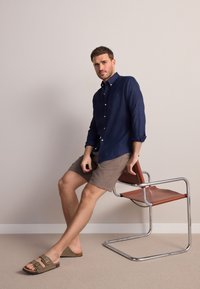 Camicia blu navy con bottoni, pantaloncini marroni e sandali taupe. Sedia con struttura in acciaio e seduta marrone. Texture lisce e design semplice.