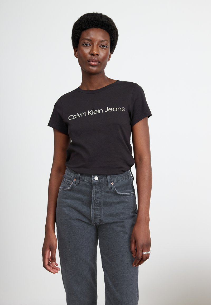 Žena s kratkom kovrčavom kosom koja nosi crnu Calvin Klein Jeans majicu i tamnosive traperice visokog struka, stoji ispred jednobojne pozadine.
