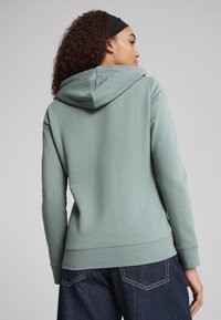 Felpa verde con cappuccio regolabile, tessuto morbido, maniche lunghe e polsini a coste, abbinata a pantaloni in denim a vestibilità aderente.