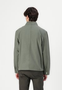 Lys grøn bomuldsjakke med spidse kraver, knapærmer og lige skæring. Syninger er synlige på bagsiden, minimalistisk design.
